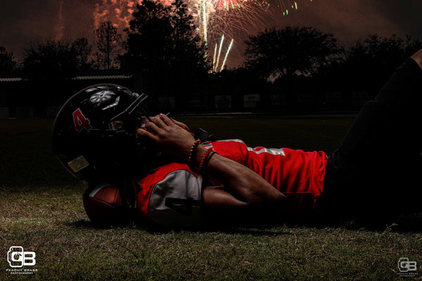 Football player laying his head on a football in grass with fireworks in the sky